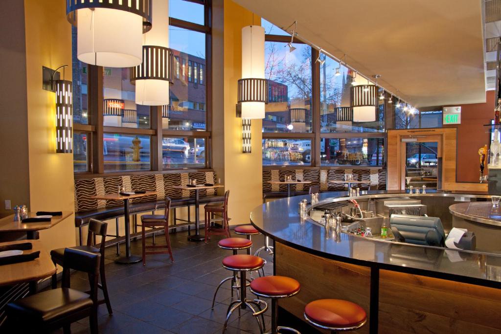 A hotel bar and lounge at the Silver Cloud Hotel Seattle Broadway featuring a curved stainless steel bar counter with orange leather stools, cylindrical drum pendant lights, wall sconces, banquette seating, and floor-to-ceiling windows overlooking Broadway in Seattle, Washington, USA.