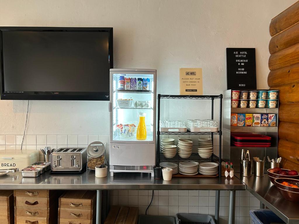 A self-serve breakfast station at the Ace Hotel Seattle featuring a mounted TV, a mini fridge stocked with juices and yogurt, wire shelving with stacked bowls and plates, a cereal display, a toaster, bread, and a bowl of fresh fruit on a stainless steel counter in Seattle, Washington, USA.