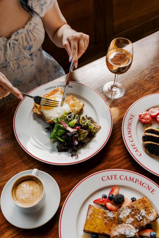 An overhead view of a woman cutting into a Café Campagne breakfast dish of poached egg over toast with mixed greens, alongside a glass of sparkling wine, a latte, sliced tomatoes, a chocolate croissant, and a plate of French toast with berries at the Inn at the Market in Seattle, Washington, USA.