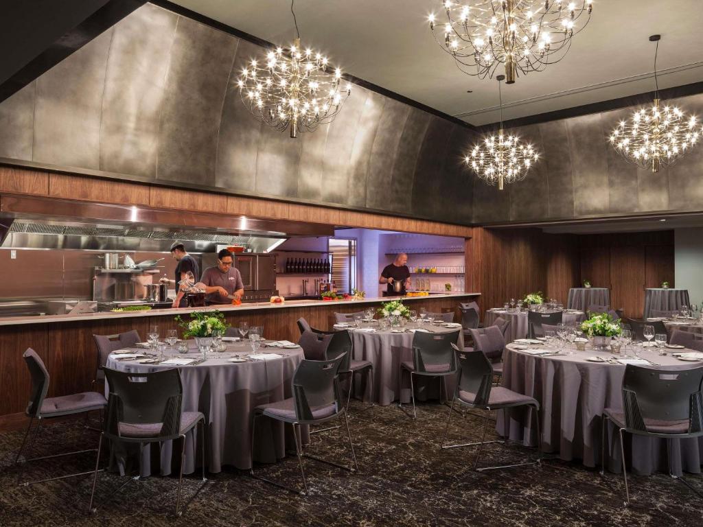 A dining area at the Ändra Hotel Seattle featuring round tables set with gray linens, white floral centerpieces, glassware, and place settings, with chefs preparing food at an open exhibition kitchen counter beneath sculptural gold starburst chandeliers in Seattle, Washington, USA.