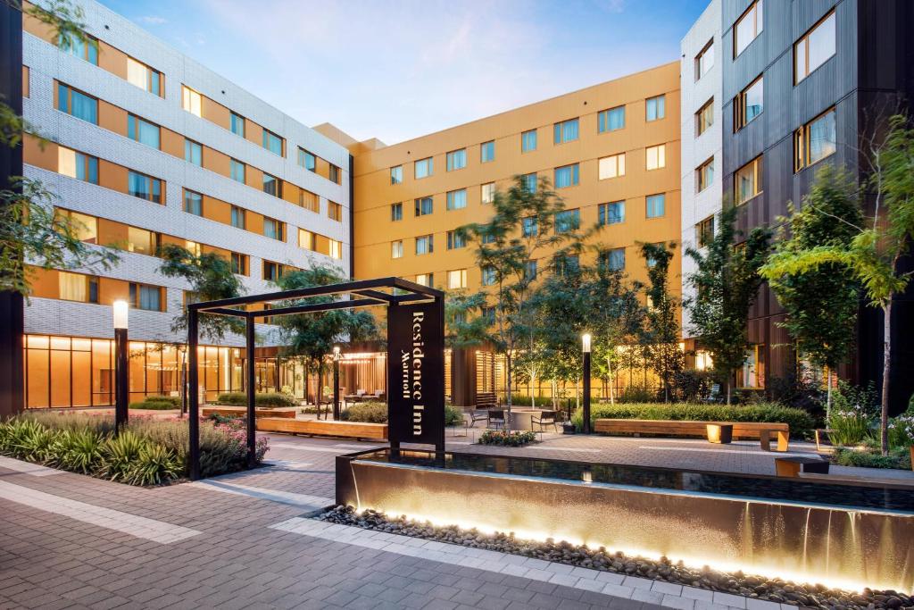 Illuminated entrance courtyard of Residence Inn by Marriott Portland Downtown at dusk, featuring a lit black signage pergola, cascading stone water feature, landscaped walkway with greenery and string lights, and multi-story tan and gray buildings with warm interior lighting in Portland, Oregon.