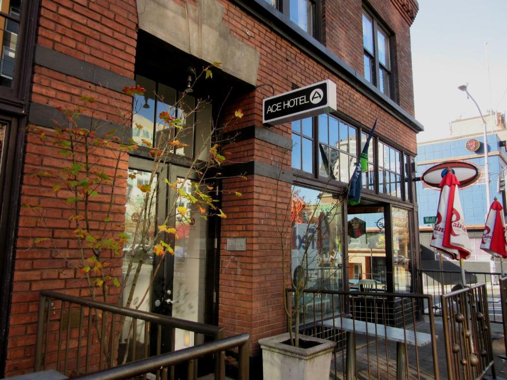 The brick exterior of the Ace Hotel Seattle featuring a black and white hanging sign above the entrance, large glass windows, wrought iron railings, potted trees with autumn leaves, and red Rainier Beer patio umbrellas on Wall Street in Seattle, Washington, USA.