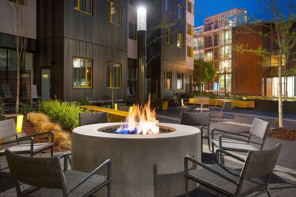 Outdoor fire pit with blue and orange flames burning in a round concrete surround, encircled by gray wicker lounge chairs on a stone patio at dusk, with tall modern apartment-style buildings, string lighting, and landscaped greenery at Residence Inn by Marriott Portland Downtown, Portland, Oregon.