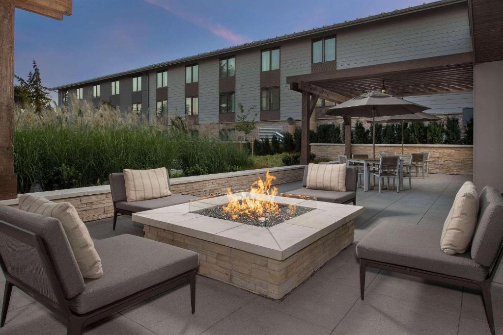 An outdoor patio at the Country Inn & Suites by Radisson, Seattle-Tacoma Airport featuring a square gas fire pit surrounded by gray cushioned lounge chairs, a covered dining area with umbrellas, and ornamental grasses in front of the hotel building at dusk in SeaTac, Washington, USA.