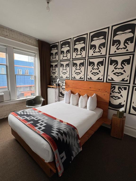 A hotel guest room at the Ace Hotel Seattle featuring a wood-plank bed with white pillows and a striped Pendleton-style blanket, a wall covered in repeating black-and-white stenciled face artwork reminiscent of Shepard Fairey street art, and a window with a city view in Seattle, Washington, USA.