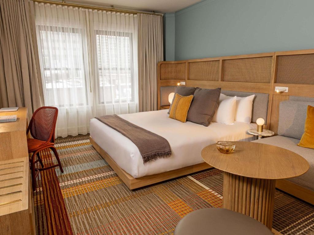 A hotel guest room at the Ändra Hotel Seattle featuring a bed with white linens and a gray throw against a woven cane headboard, mustard and gray accent pillows, a round wood side table, a red cane chair at a desk, and sheer curtains over the window in Seattle, Washington, USA.