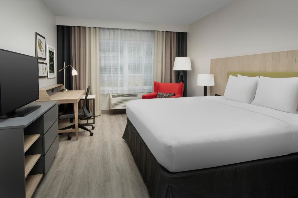 A hotel guest room at the Country Inn & Suites by Radisson, Seattle-Tacoma Airport featuring a king bed with crisp white linens and a black skirt, a red accent armchair, a wood desk with an office chair, a wall-mounted flat-screen TV, and sheer curtains over the window in SeaTac, Washington, USA.