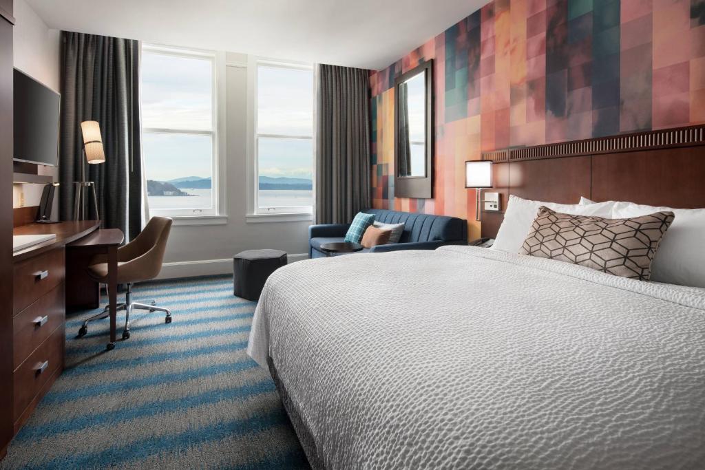 A hotel guest room at the Courtyard by Marriott Seattle Downtown Pioneer Square featuring a king bed with white linens against a colorful patchwork wallpaper accent wall, a navy sofa, a wood desk with an office chair, and a water view through large windows in Seattle, Washington, USA.