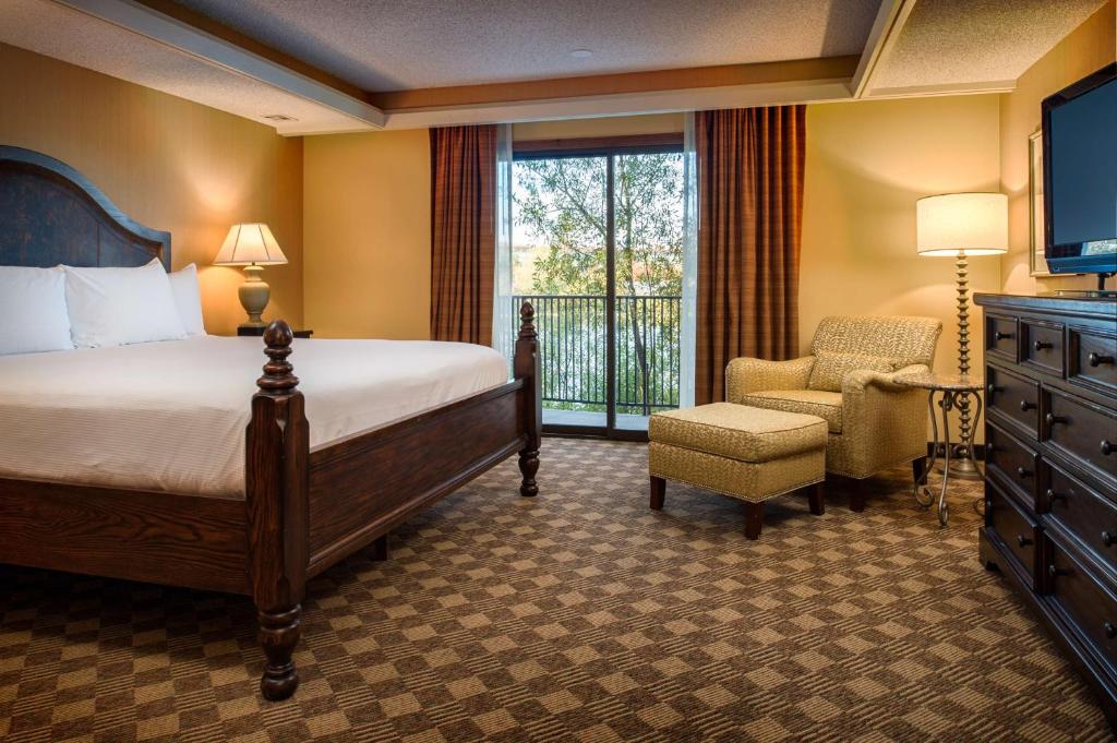 A hotel guest room at the DoubleTree by Hilton Hotel Seattle Airport featuring a dark wood four-poster bed with white linens, a matching wood dresser and TV stand, a cushioned armchair with ottoman, and a sliding glass door leading to a balcony in SeaTac, Washington, USA.