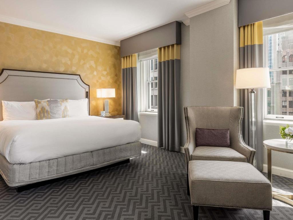 A hotel guest room at the Fairmont Olympic Hotel featuring a king bed with white linens and a gray upholstered headboard against a gold patterned accent wall, a wingback armchair with an ottoman, a round marble side table, and a city view through large windows in Seattle, Washington, USA.