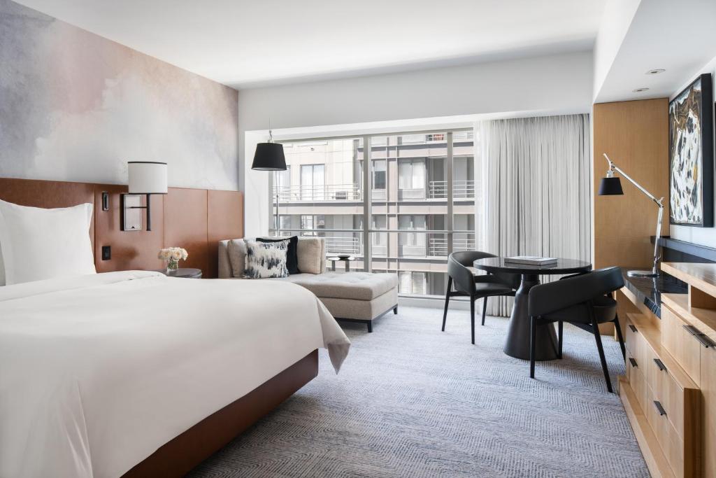 A hotel guest room at the Four Seasons Hotel Seattle featuring a king bed with a brown leather headboard, an abstract watercolor-style wall mural, a gray chaise lounge, a round black dining table with chairs, and floor-to-ceiling windows overlooking the city in Seattle, Washington, USA.