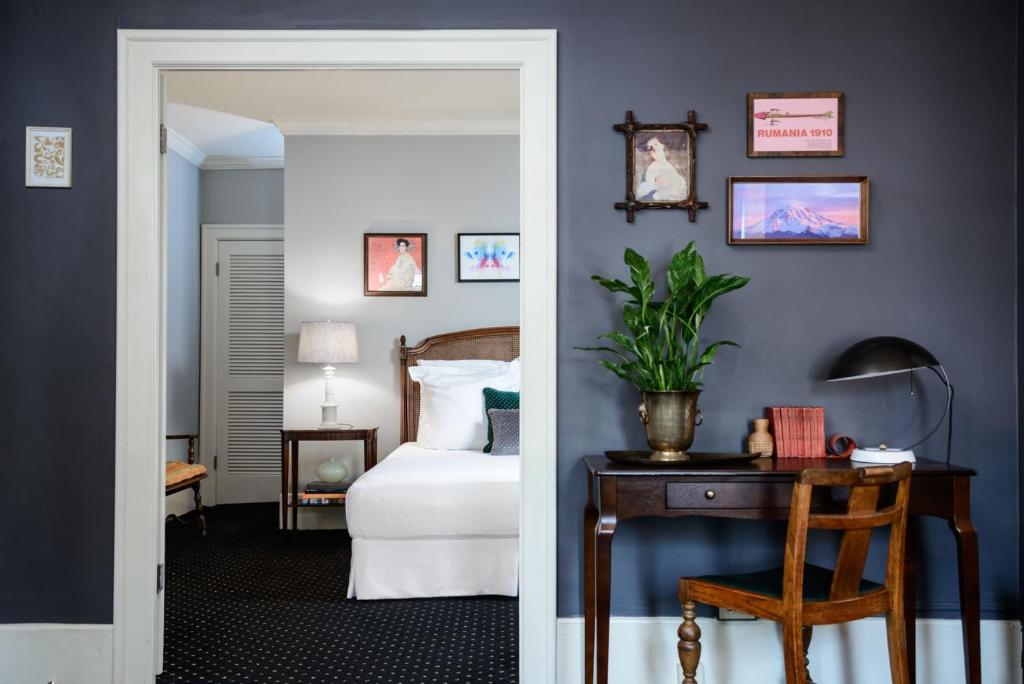 A hotel suite at the Hotel Sorrento featuring a navy blue accent wall with a curated gallery of framed art, a brass desk lamp, an antique wood writing desk and chair, a potted plant, and a view into an adjoining bedroom with a cane headboard and white linens in Seattle, Washington, USA.