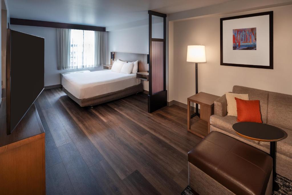 A modern hotel guest room at the Hyatt Place Seattle Downtown featuring a queen bed with crisp white linens, dark wood-look flooring, a brown sectional sofa with an orange accent pillow, a lit floor lamp, framed abstract wall art, and a large flat-screen TV in Seattle, Washington, USA.