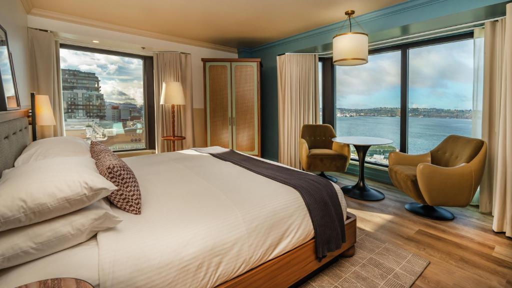 A hotel guest room at the Inn at the Market featuring a bed with white linens and a striped throw, a rattan wardrobe, two mustard-colored swivel armchairs around a round table, a pendant light, and floor-to-ceiling windows overlooking Elliott Bay in Seattle, Washington, USA.
