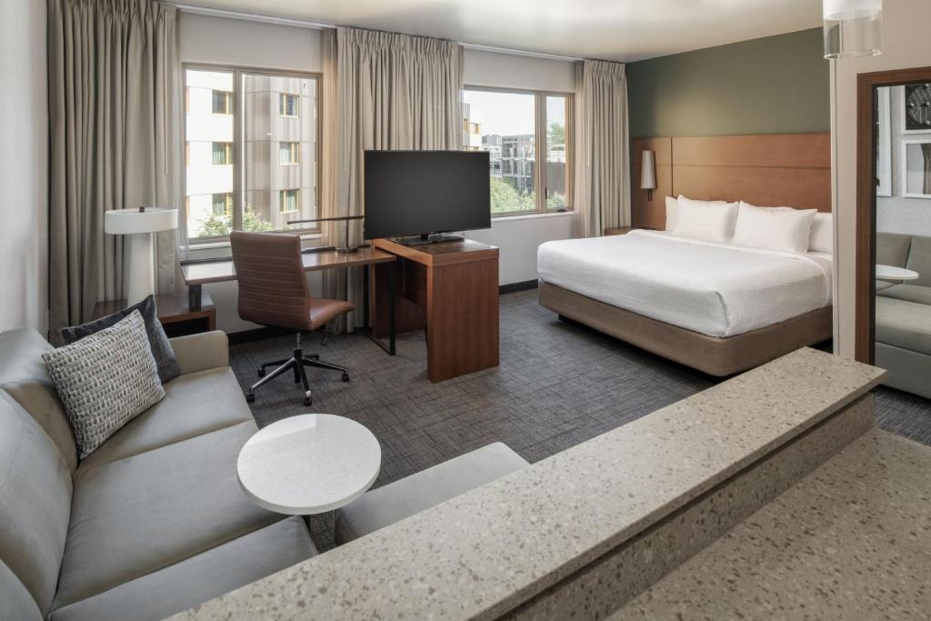 Modern hotel guest room featuring a king bed with white linens and padded wood headboard, a gray sofa with striped pillows, round side table, wood desk with leather chair, mounted flat-screen TV, and windows overlooking the city at Residence Inn by Marriott Portland Downtown, Portland, Oregon.