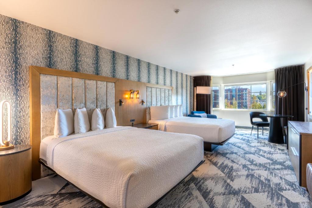 A hotel guest room at the Silver Cloud Hotel Seattle Broadway featuring two queen beds with white linens against a wood-framed upholstered headboard and a textured blue and white accent wall, a round wood nightstand, a black dining table with chairs, and a city view window in Seattle, Washington, USA.