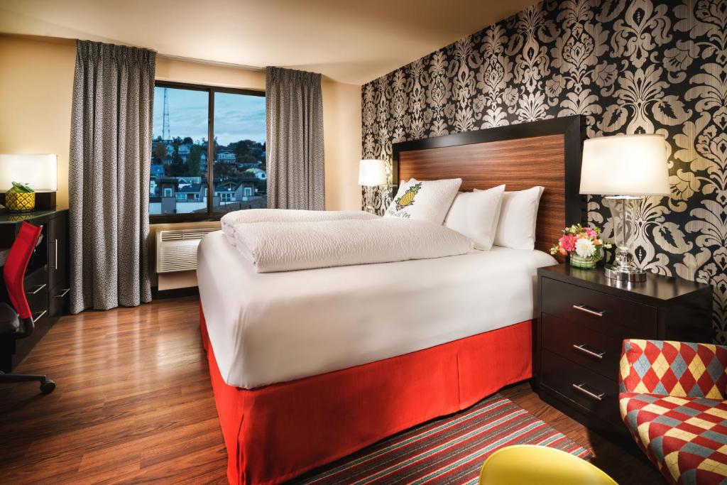 A hotel guest room at Staypineapple, The Maxwell Hotel Seattle Center featuring a bed with white linens, a red skirt, and pineapple-embroidered pillows against a black and silver damask accent wall, a wood headboard, a red desk chair, and a harlequin-patterned armchair in Seattle, Washington, USA.