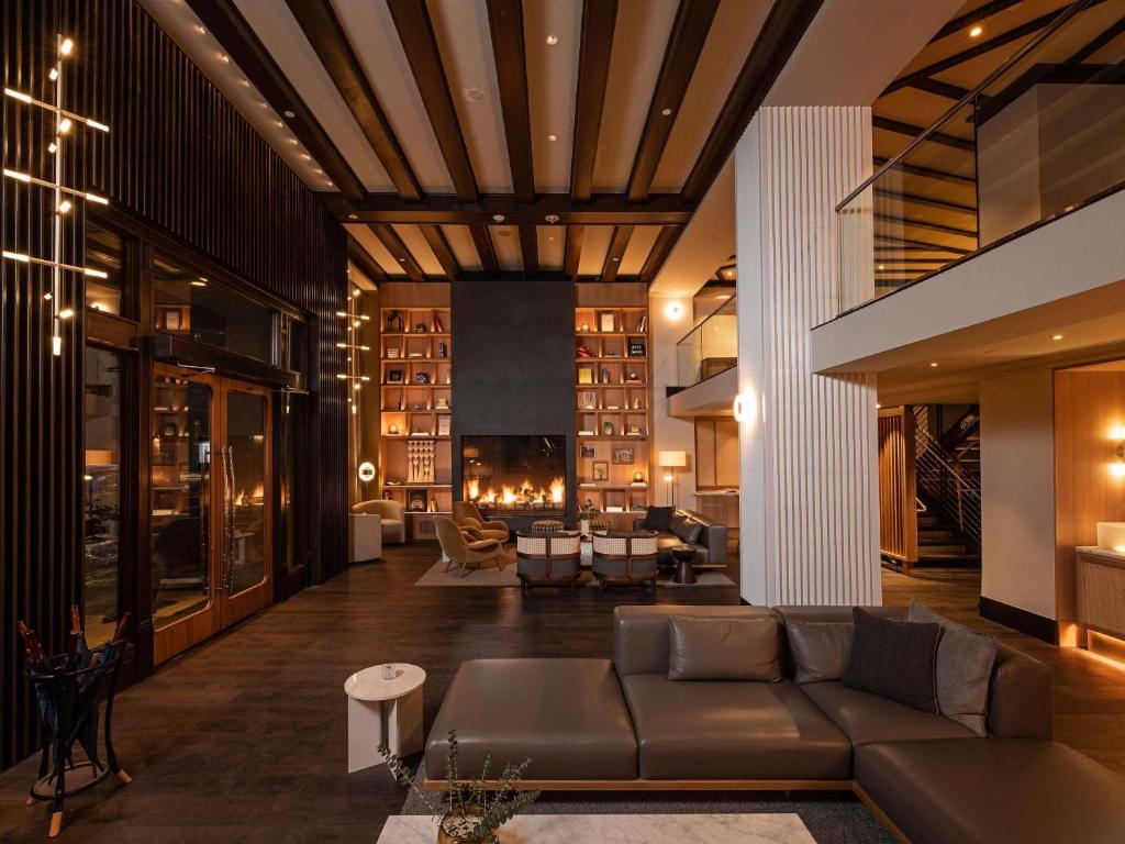 A hotel lobby lounge at the Ändra Hotel Seattle featuring a dark leather sectional sofa, a built-in bookshelf surrounding a lit fireplace, wood-beamed ceilings, cane accent chairs, and a mezzanine level with a glass railing visible above in Seattle, Washington, USA.