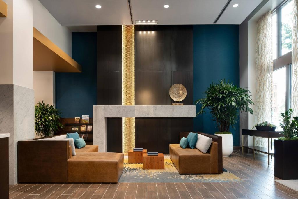 A hotel lobby lounge at the Courtyard by Marriott Seattle Downtown Pioneer Square featuring tan leather sofas with teal and white throw pillows, a wood coffee table, a fireplace with a decorative gold disc above the mantel, potted palms, and a teal accent wall in Seattle, Washington, USA.