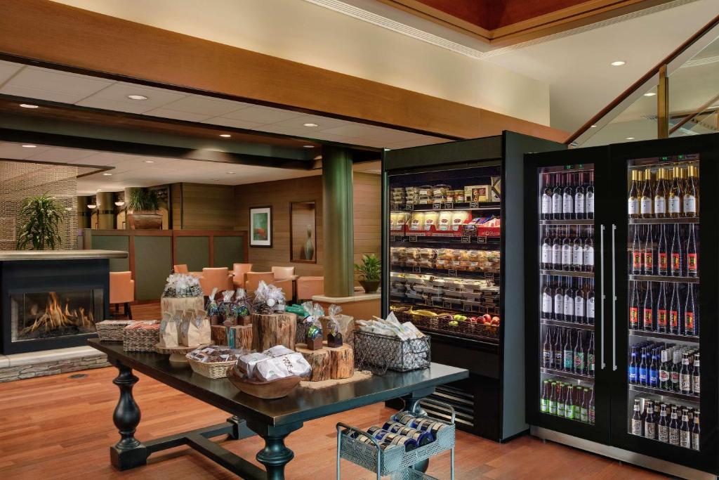 A hotel marketplace at the DoubleTree by Hilton Hotel Seattle Airport featuring a wood display table with packaged snacks and gift baskets, a refrigerated case stocked with fresh food, glass-front coolers filled with wine and beer, and a lit stone fireplace nearby in SeaTac, Washington, USA.