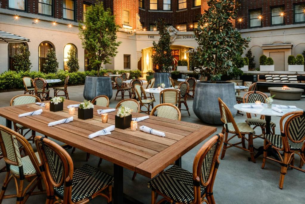An outdoor courtyard at the Hotel Sorrento in the evening featuring a long wooden dining table set with candles and floral centerpieces, woven bistro chairs, potted topiary trees, string lights, and a lit fire pit with striped lounge seating in Seattle, Washington, USA.