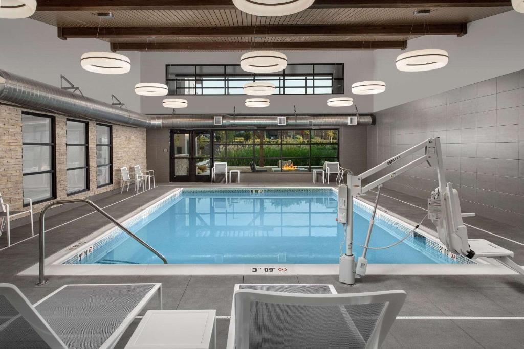 An indoor swimming pool at the Country Inn & Suites by Radisson, Seattle-Tacoma Airport featuring a rectangular pool with a pool lift, poolside lounge chairs, exposed wood-beam ceiling, round pendant lighting, and a view of an outdoor fire pit through the windows in SeaTac, Washington, USA.