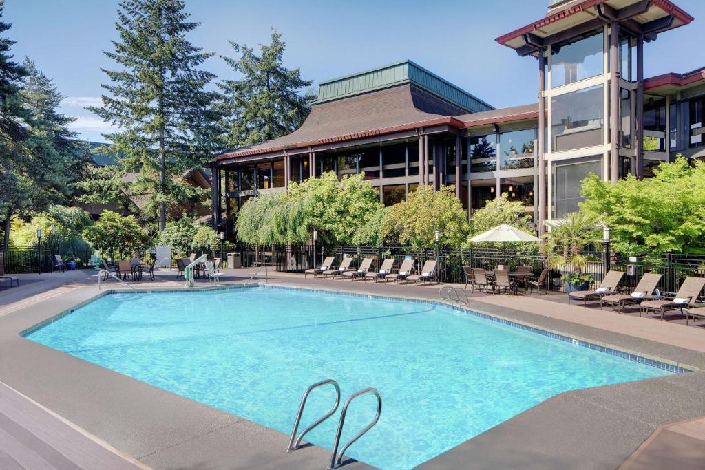An outdoor swimming pool at the DoubleTree by Hilton Hotel Seattle Airport featuring a rectangular pool with a metal ladder, poolside lounge chairs, a dining table with an umbrella, tall evergreen trees, and the angular timber-roofed hotel building in the background in SeaTac, Washington, USA.