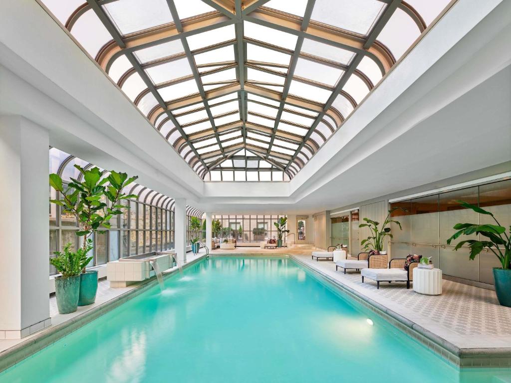 An indoor swimming pool at the Fairmont Olympic Hotel featuring a long rectangular pool beneath an arched glass skylight, white lounge chairs and chaises along the poolside, potted palm and bird of paradise plants, and floor-to-ceiling windows in Seattle, Washington, USA.