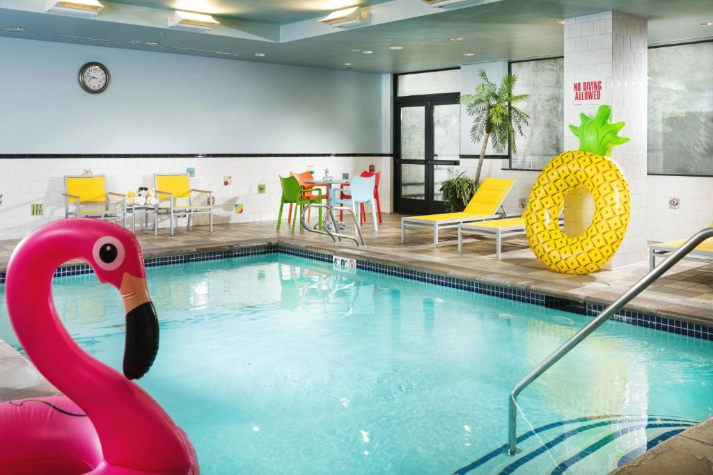 An indoor swimming pool at Staypineapple, The Maxwell Hotel Seattle Center featuring a pink inflatable flamingo float and a yellow inflatable pineapple ring in the water, yellow poolside lounge chairs, a decorative palm tree, and colorful mismatched dining chairs in Seattle, Washington, USA.