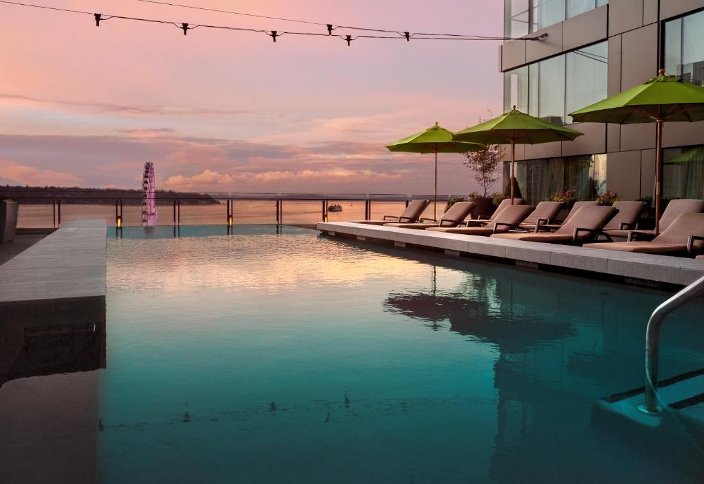 A rooftop infinity pool at the Four Seasons Hotel Seattle overlooking Elliott Bay at sunset, with tan lounge chairs and green umbrellas along the poolside, the illuminated Seattle Great Wheel visible on the waterfront, and a pink and orange sky reflected in the water in Seattle, Washington, USA.