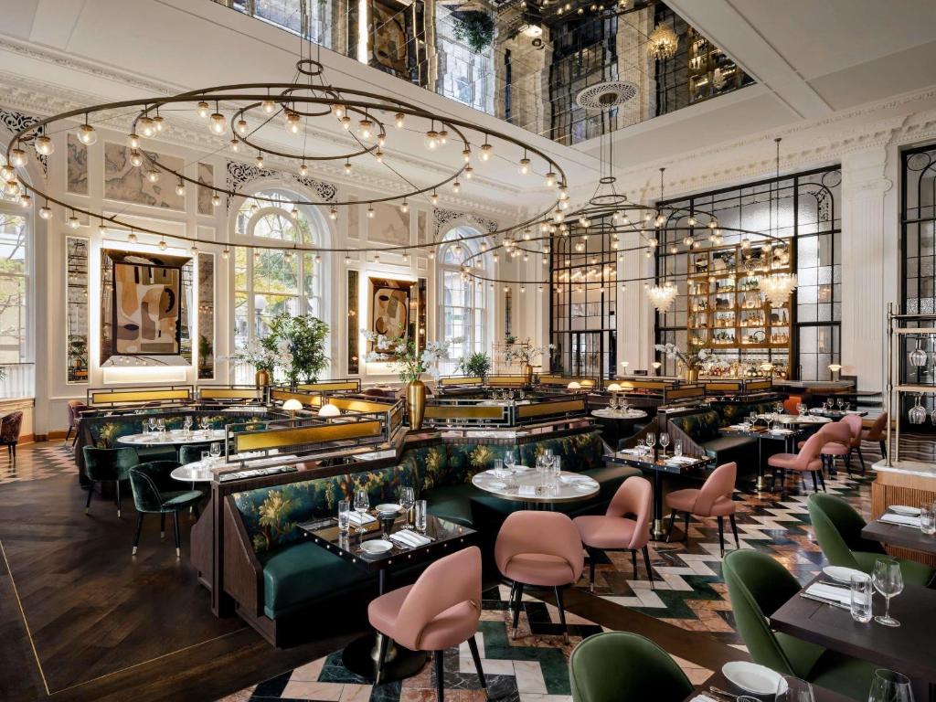 An elegant restaurant dining room at the Fairmont Olympic Hotel featuring pink and green upholstered chairs, marble checkerboard flooring, ornate white pillars, arched windows, a mirrored ceiling, and circular string-light chandeliers hanging above the tables in Seattle, Washington, USA.