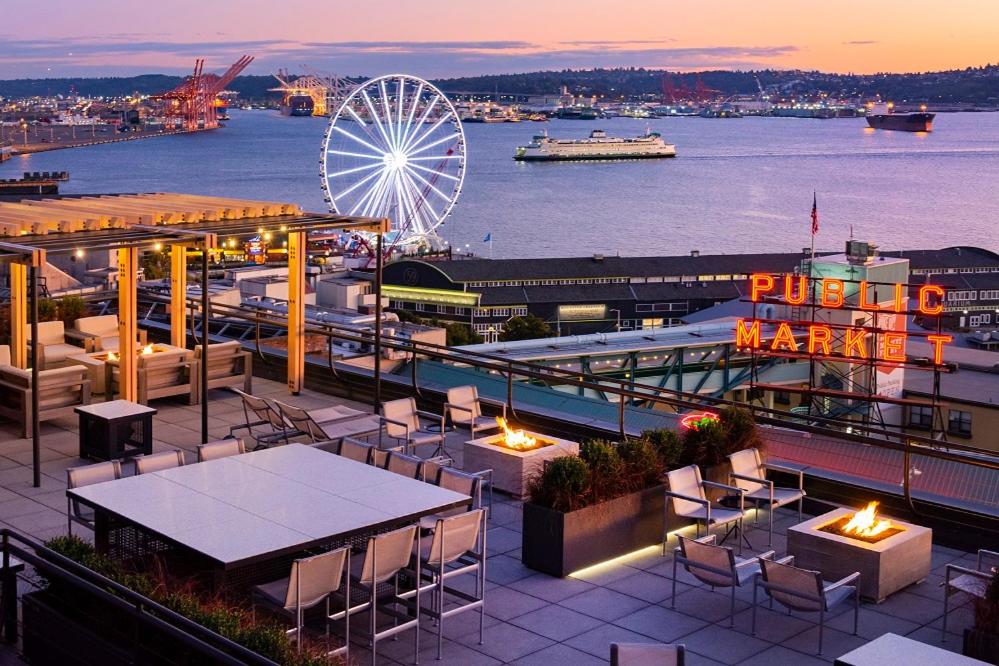 A rooftop lounge at the Inn at the Market at sunset featuring lounge seating, fire pit tables, and a wooden pergola overlooking Elliott Bay, with the illuminated Seattle Great Wheel and the neon Pike Place Public Market sign visible against a pink sky in Seattle, Washington, USA.