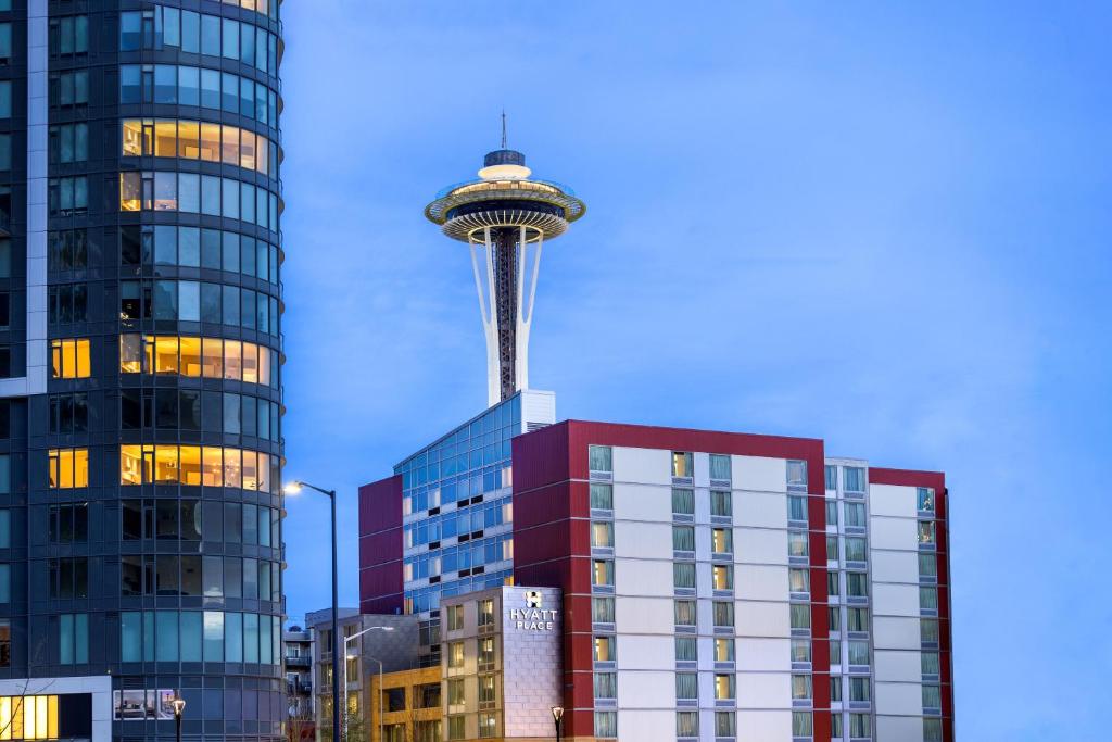 The exterior of the Hyatt Place Seattle Downtown hotel rises in maroon and cream tones at dusk, with the brightly illuminated Space Needle towering directly behind it against a clear blue sky, and a tall glass high-rise apartment building visible to the left in Seattle, Washington, USA.