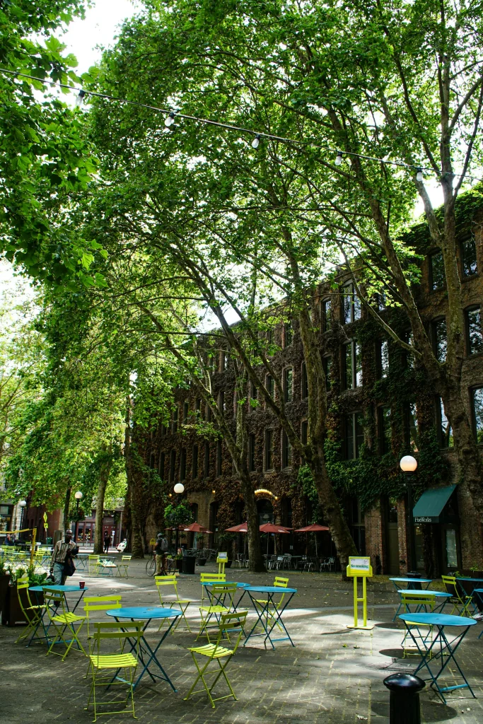 Occidental Square in Seattle’s historic Pioneer Square neighborhood, featuring colorful outdoor tables and chairs beneath leafy trees beside ivy-covered brick buildings.