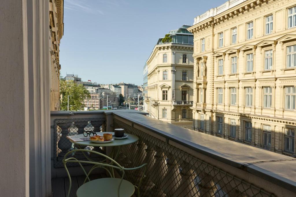 Sunny private hotel balcony at Cloud One Wien-Staatsoper with a pale green bistro table set for breakfast with pastries, coffee, and reading glasses, behind an ornate stone balustrade overlooking grand cream Historicist buildings and a leafy boulevard near the State Opera in Vienna, Austria.