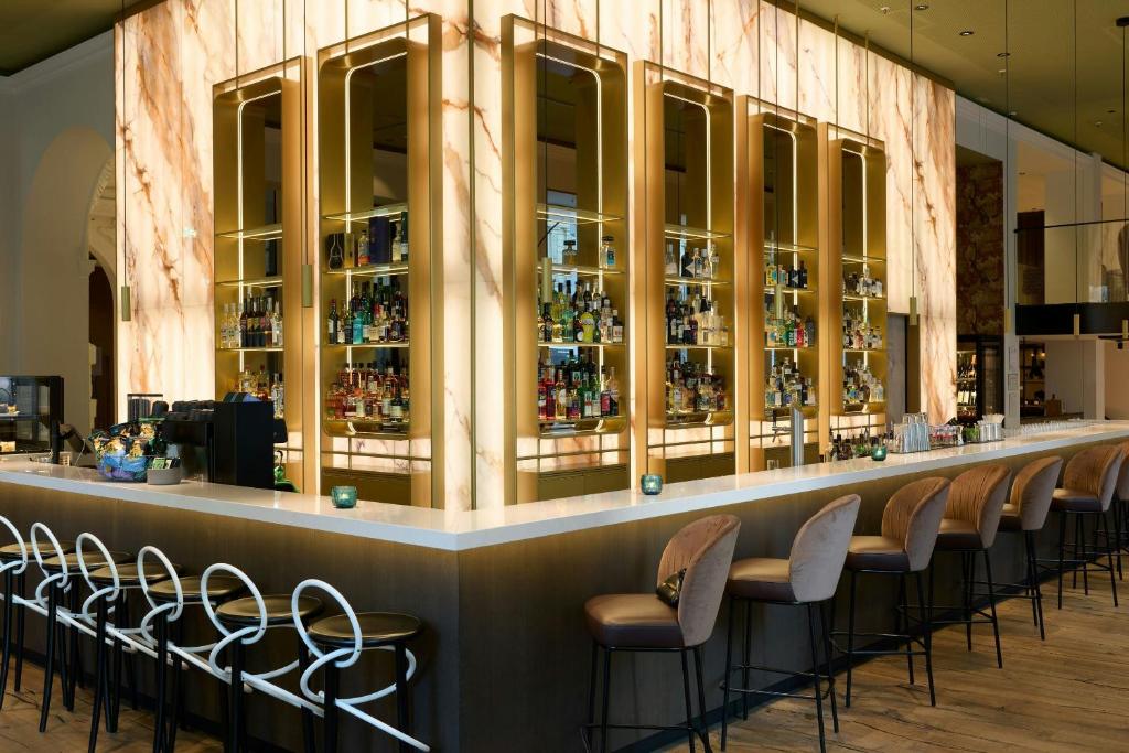 Elegant hotel bar at Cloud One Wien-Staatsoper with a long white counter, backlit onyx marble walls, and illuminated brass-framed shelves stocked with spirits, lined with blush velvet bar stools and white looped metal stools on a light wood floor near the State Opera in Vienna, Austria.
