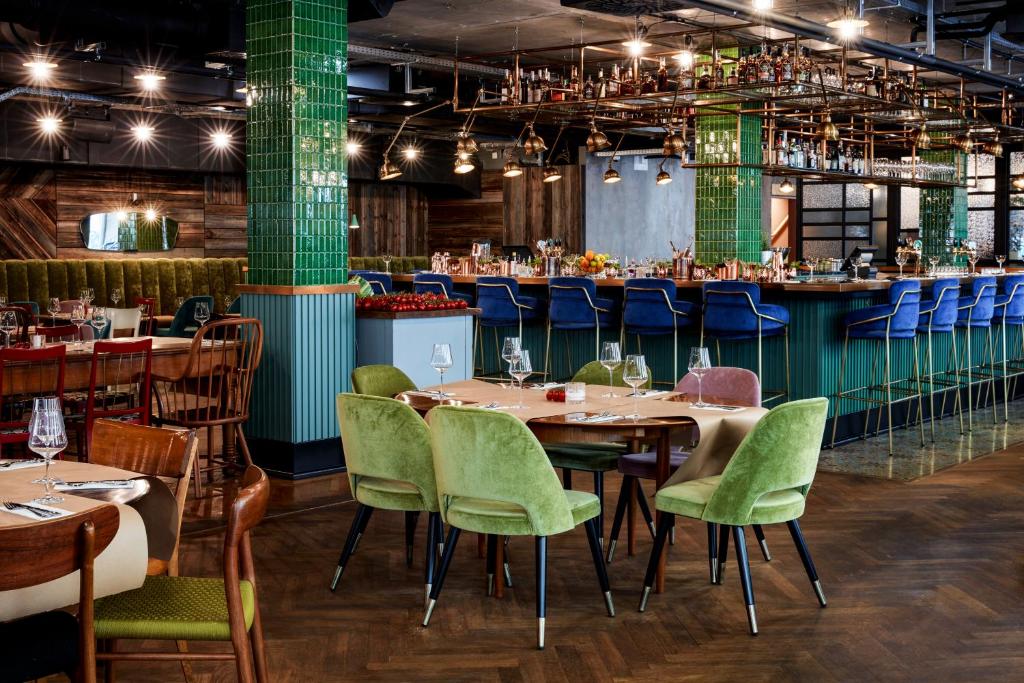 Restaurant and bar at Max Brown Hotel 7th District with emerald green tiled columns, a copper hanging shelf of spirits above the bar, cobalt velvet bar stools, lime green velvet dining chairs around tables set with wine glasses, wood-paneled walls, and dark floors in Vienna, Austria.