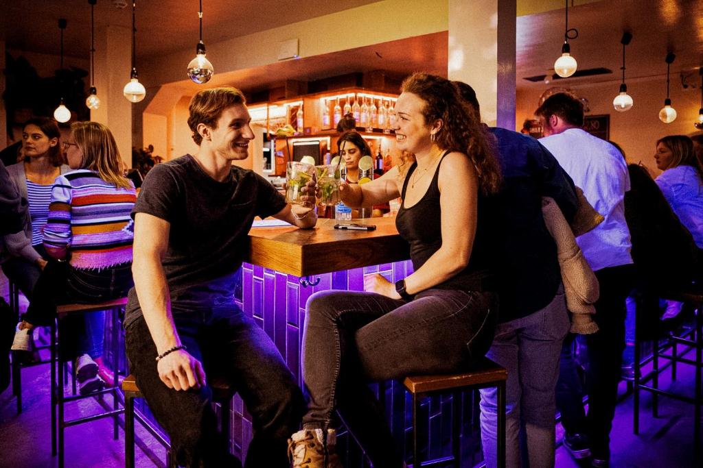 Lively hostel bar at Wombat’s City Hostel Vienna Naschmarkt where smiling young travelers clink mojito glasses at a wooden counter with purple-lit tiles, other guests chatting on bar stools, a bartender beside backlit shelves of spirits, and hanging bulb pendant lights in Vienna, Austria.