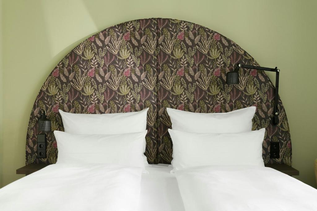 Cozy Cloud One Wien-Staatsoper guest room double bed with crisp white linens and four fluffy pillows set against an arched upholstered headboard in a dark botanical print of green, pink, and gold leaves, framed by sage green walls and matte black swing-arm reading lamps in Vienna, Austria.