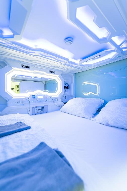 Inside a double sleep capsule at Space Home Apartment - City Center, where glowing blue LED lighting outlines a spaceship-style ceiling and porthole windows above a white bed with two pillows, folded gray towels, a control panel, headphones, and a small in-pod safe in Vienna, Austria.