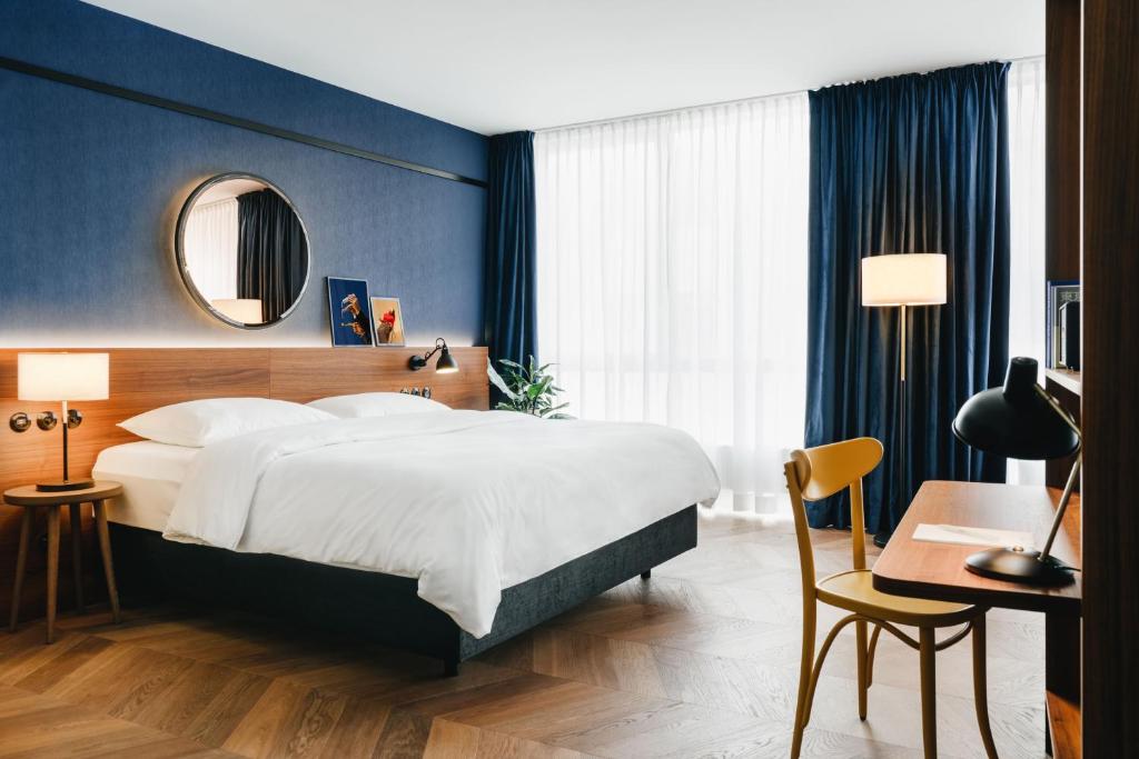 Hotel Gilbert guest bedroom with a king bed in crisp white linens, a navy fabric accent wall with a round mirror and framed bird portrait art, a walnut headboard with reading lights, blue velvet curtains, herringbone wood floors, and a writing desk with a bentwood chair in Vienna, Austria.