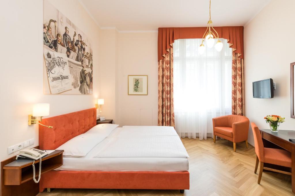 Hotel Johann Strauss guest room with a double bed and tufted orange upholstered headboard, a large mural of Johann Strauss’s Kaiser-Walzer sheet music and musicians, orange swagged curtains, a brass chandelier, a matching armchair and desk, and herringbone wood floors in Vienna, Austria.