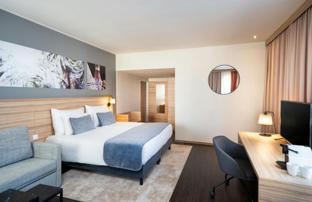 Modern guest room at Leonardo Hotel Vienna Hauptbahnhof with a king bed dressed in white linens and a blue throw, a light wood headboard beneath botanical and city art prints, a gray sofa, a round wall mirror, a work desk with coffee maker and TV, and blush curtains in Vienna, Austria.