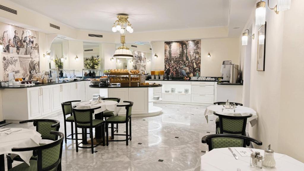 Hotel Johann Strauss breakfast room with a buffet of fresh breads, pastries, and juices on white cabinetry, round tables with white cloths and green upholstered chairs, polished white marble floors, an ornate brass chandelier, and murals of Viennese waltz-era musicians in Vienna, Austria.