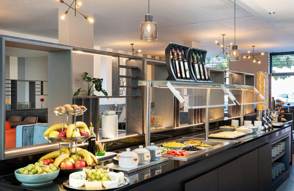Breakfast buffet at Leonardo Hotel Vienna Hauptbahnhof with tiered stands of bananas, apples, and kiwis, a bowl of green grapes, a cheese platter, sliced vegetables, olives, and covered jars on a long black counter beneath glass sneeze guards and modern pendant lights in Vienna, Austria.