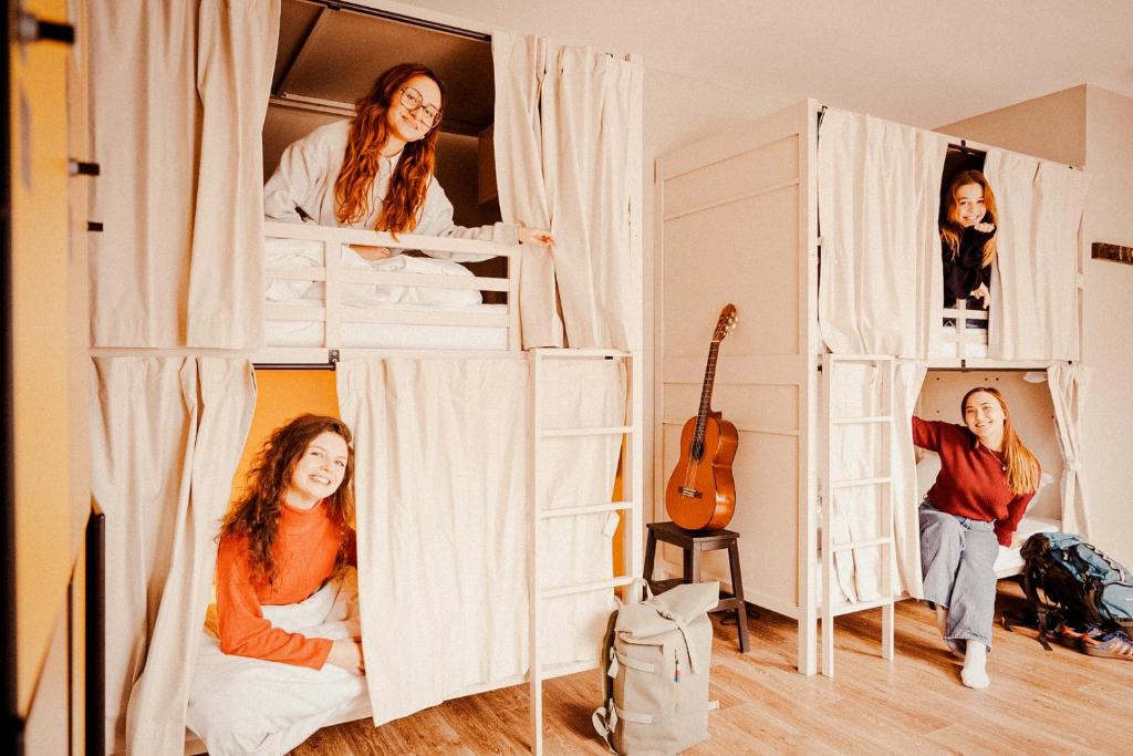 Dorm room at Wombat’s City Hostel Vienna Naschmarkt with cream-curtained wooden bunk-bed pods and ladders, where four smiling young women travelers peek out from their beds, with an acoustic guitar on a stool, backpacks on the light wood floor, and orange accent walls in Vienna, Austria.