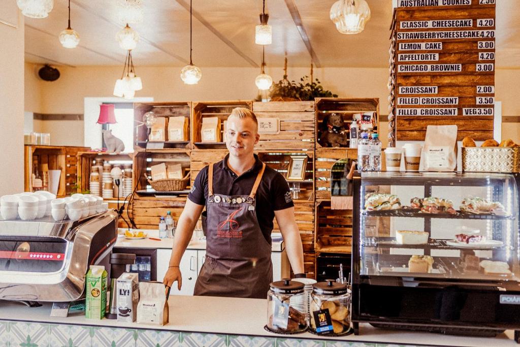 Smiling young barista in a branded apron behind the café counter at Wombat’s City Hostel Vienna Naschmarkt, with an espresso machine, a pastry case of sandwiches and cakes, wooden crate shelving of coffee bags, vintage glass pendant lights, and a wooden menu board of cakes in Vienna, Austria.