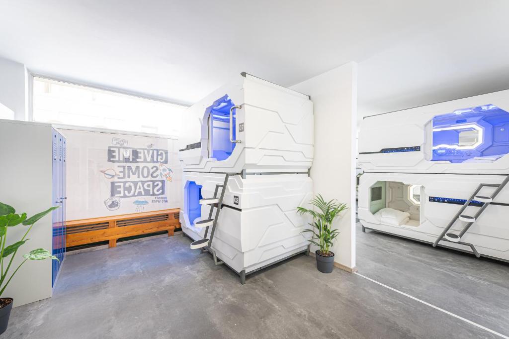 Futuristic white sleep pods stacked two high with glowing blue windows, numbered doors, and metal step ladders at Space Home Apartment - City Center, next to tall blue lockers, two potted palms, and a playful wall mural reading Give Me Some Space above a wooden bench in Vienna, Austria.
