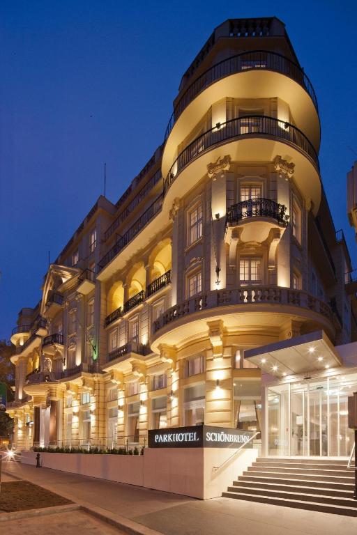 Illuminated Historicist facade of Austria Trend Parkhotel Schönbrunn at twilight, with a rounded corner tower, wrought-iron balconies, ornate cream stonework glowing against a deep blue sky, and a sleek modern glass entrance pavilion with stairs beside the hotel sign in Vienna, Austria.