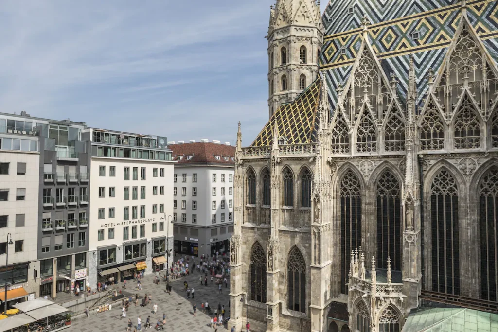 An elevated view of Boutique Hotel am Stephansplatz facing St. Stephen’s Cathedral, its Gothic spires, tracery windows, and colorful zigzag tiled roof towering above crowds strolling the pedestrian Stephansplatz square on a sunny summer day in the historic center of Vienna, Austria.